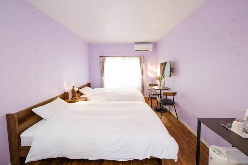 自然光が差し込む薄紫色の客室。シンプルな家具とベッド付き