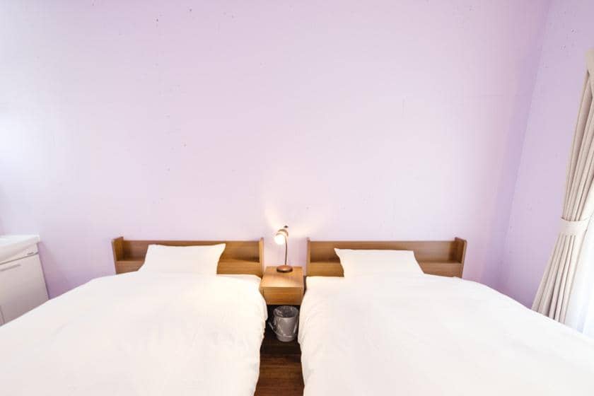  薄紫色の壁に並んだ2つのシングルベッド。間には木製のナイトテーブル