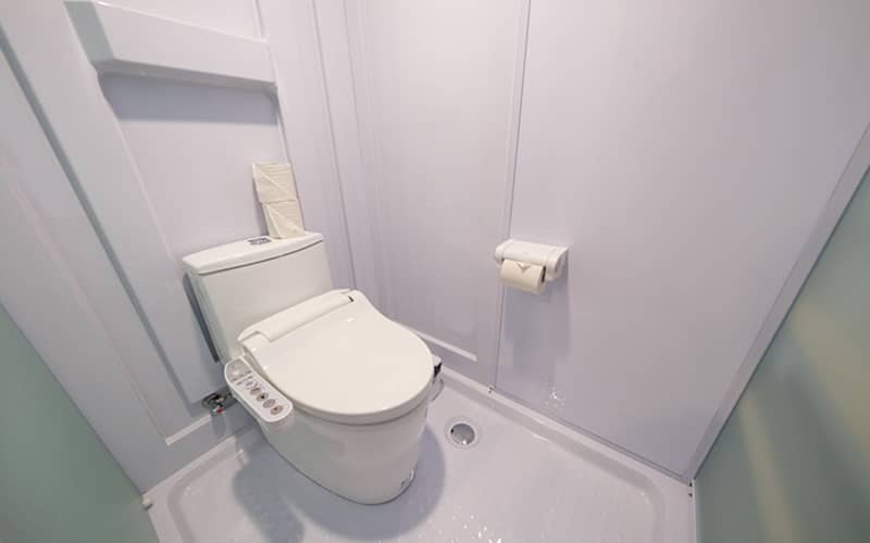清潔感のある白い温水洗浄便座付きのトイレ。壁面にはトイレットペーパーホルダーが設置され、予備のペーパーが便器背面に置かれています。床面は水に強い素材で、全体的に白で統一されたコンパクトなサニタリールームの様子です。