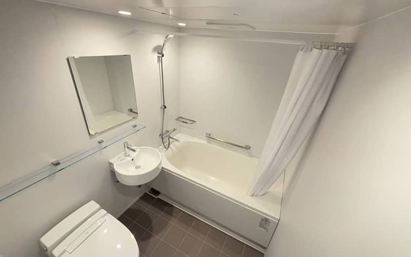 浴槽、洗面台、トイレが一体となった3点式ユニットバスの全景。左側にトイレと洗面台、正面に浴槽があり、シャワーカーテンが右側に寄せられています。壁面には大きな鏡があり、全体が白で統一された機能的な配置です。